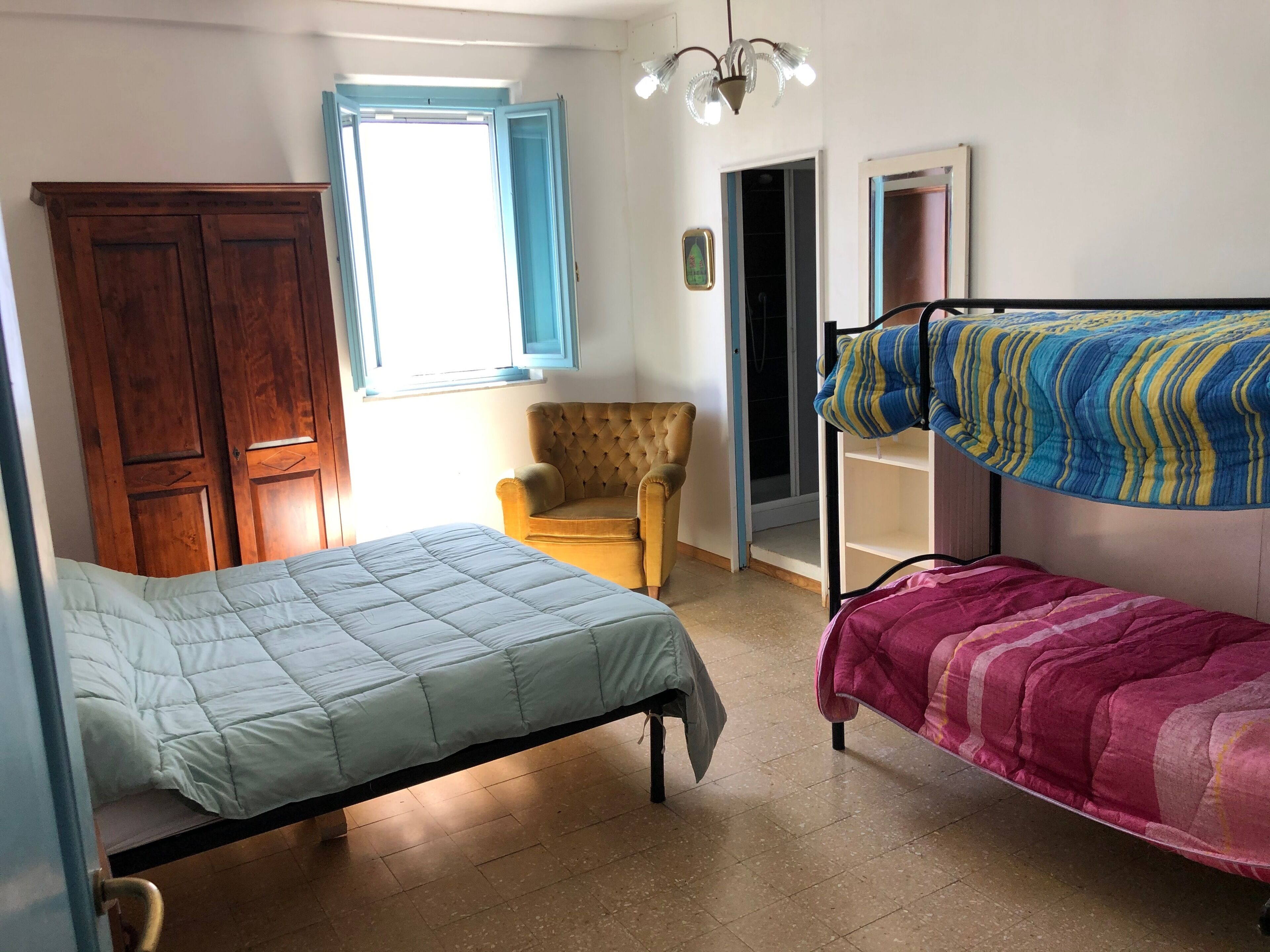 Ein Zimmer mit zwei Betten, einem Holzschrank, einem gelben Sessel und einem Fenster mit blauen Rollläden.
