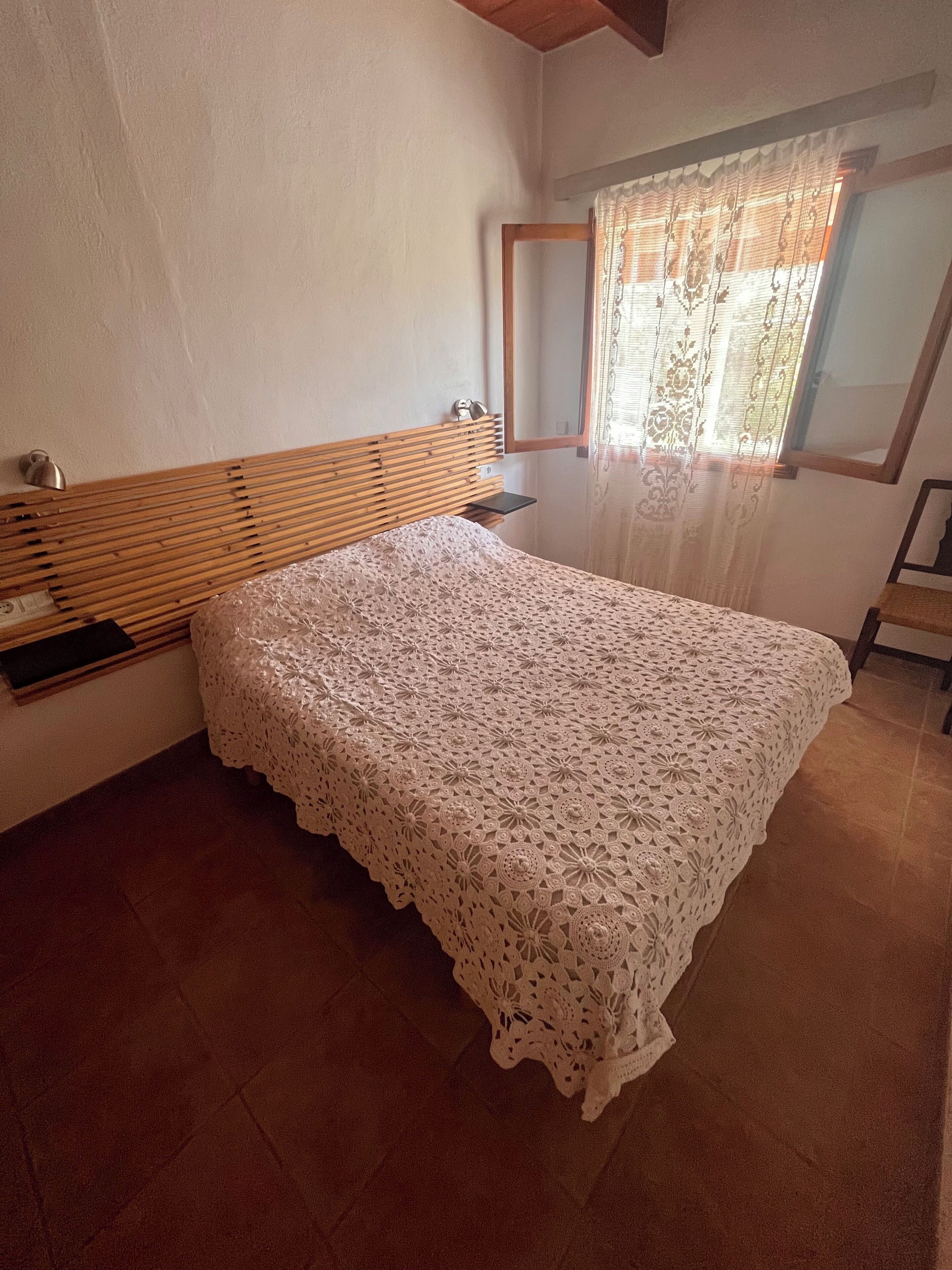 A bedroom with a wooden headboard, a lace bedspread, a bedside table, and a window with sheer curtains.