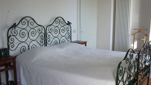Una cama con estructura metálica y una colcha blanca en una habitación con suelo de baldosas y una silla de madera.