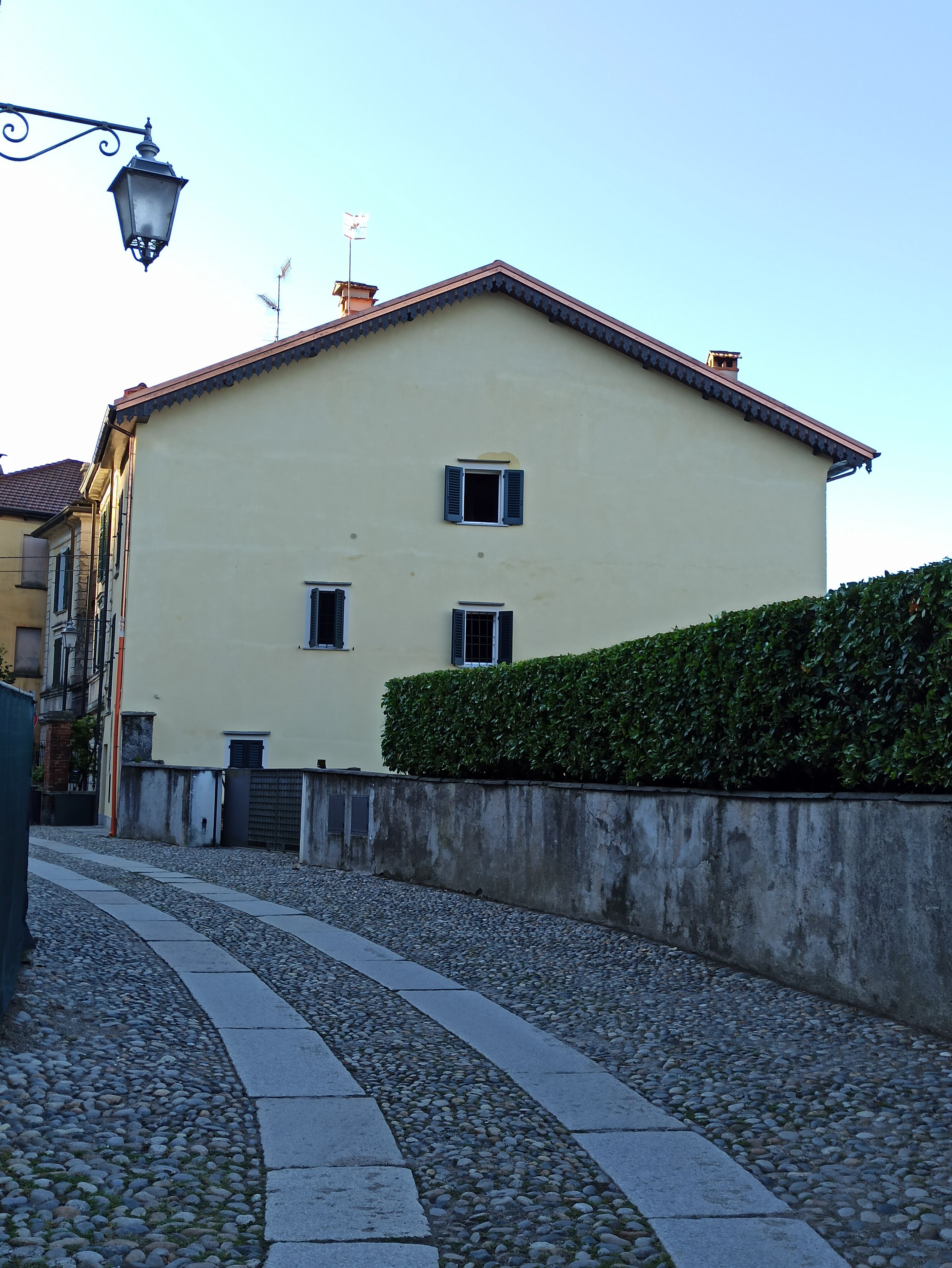 A building with a light yellow facade, a red roof, and a small window with shutters. There is a hedge in front of the building, and a cobblestone path leading to the entrance.