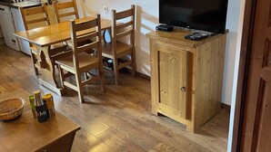 Un espacio habitable compacto con una mesa de comedor de madera, sillas, un televisor sobre un mueble de madera, una zona de cocina con microondas y una lámpara fijada a la pared.