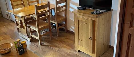 Un espacio habitable compacto con una mesa de comedor de madera, sillas, un televisor sobre un mueble de madera, una zona de cocina con microondas y una lámpara fijada a la pared.