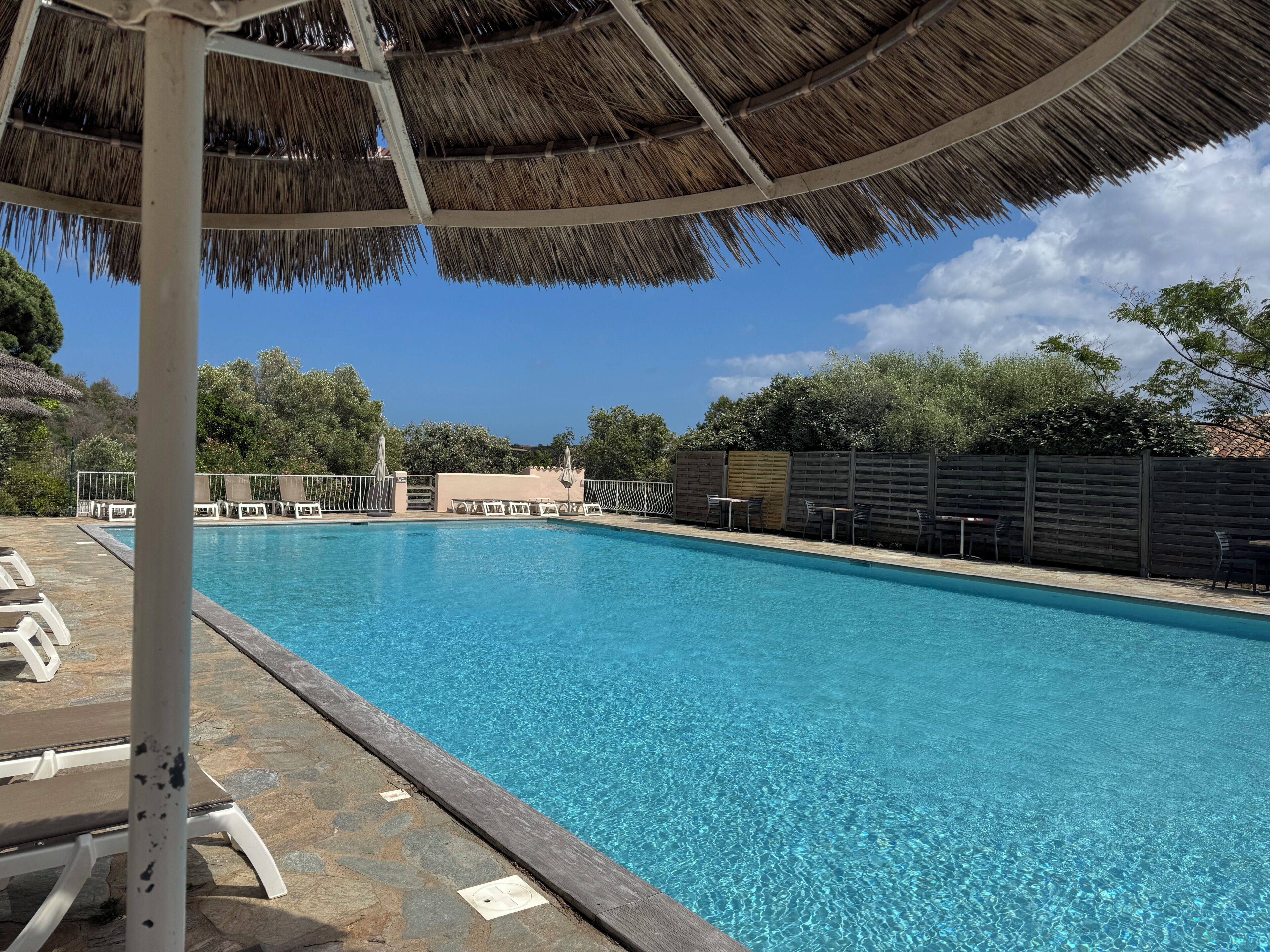Une piscine entourée d’un sol en pierre et d’une clôture en bois, avec des chaises longues et des parasols à proximité.