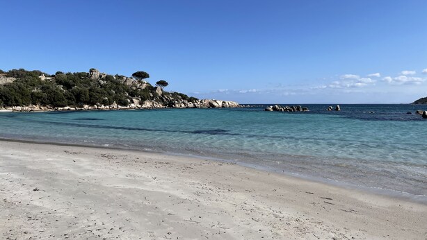 Una playa con agua turquesa cristalina, una orilla de arena y colinas rocosas al fondo.