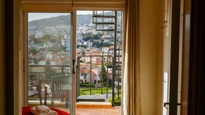 Une chambre avec un lit, un ventilateur de plafond, une vue sur le paysage urbain à travers une porte vitrée et un balcon avec une balustrade.