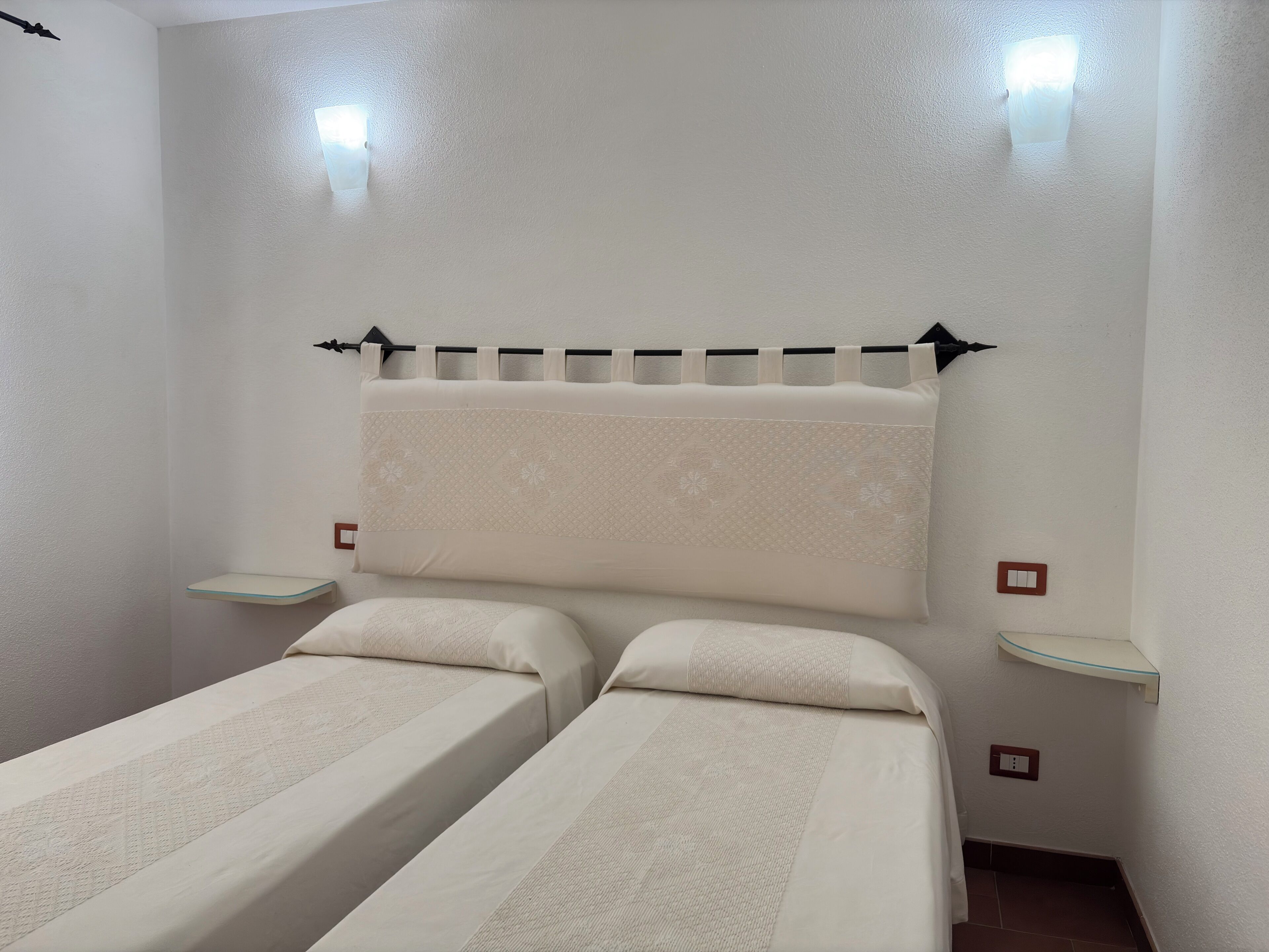 Une chambre avec deux lits, une tête de lit, des tables de chevet et des lampes fixées au mur.
