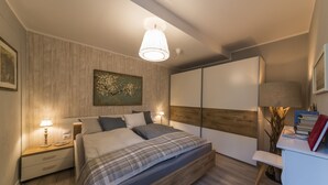 Een moderne slaapkamer met een houten hoofdbord, een groot bed met een geruite plaid, een nachtkastje met een lamp, en een bureau met boeken en een vaas.