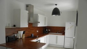 A compact kitchen with white cabinets, a wooden countertop, a black hanging light, and a refrigerator.