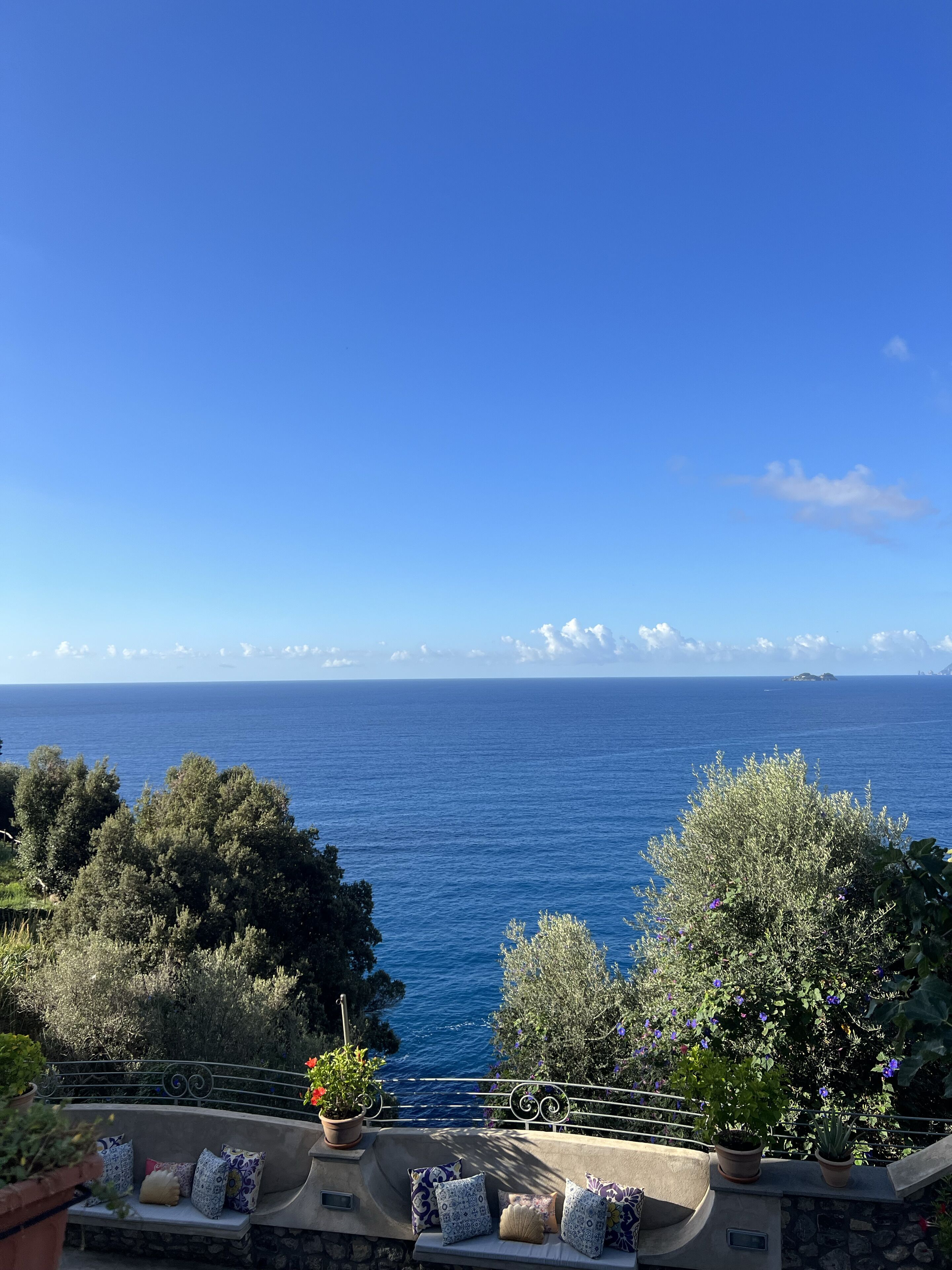 A balcony with a stone wall, a cushioned bench, and potted plants overlooking a clear blue sea with distant islands.