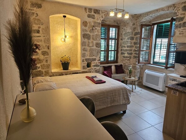 A cozy room with a bed, a small table, a potted plant, and a window with shutters.