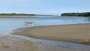 A calm beach with a boat anchored near the shore, surrounded by a clear sky and distant hills.