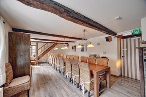 Zona pranzo con un lungo tavolo in legno e sedie in vimini, travi a vista al soffitto e una scala che conduce a un'altra stanza.