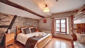 Quarto com parede de pedra, vigas de madeira, cama com almofadas, mesa de cabeceira, janela com cortinas e uma cama beliche.