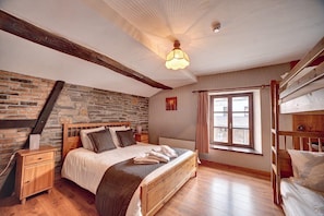 Una camera da letto con parete in pietra, travi in legno, un letto con cuscini, un comodino, una finestra con tende e un letto a castello.