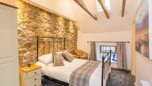 A bedroom with a stone wall, a bed with a metal frame, a wooden nightstand, a wicker chair, and a window with curtains. - Rural chic - The Hayloft others style and comfort with a rustic charm. (Balerno)
