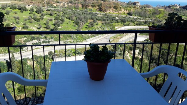 A balcony with a table and chairs, overlooking a road and hilly landscape with a view of the sea. - German house at the Borgo (Badolato)