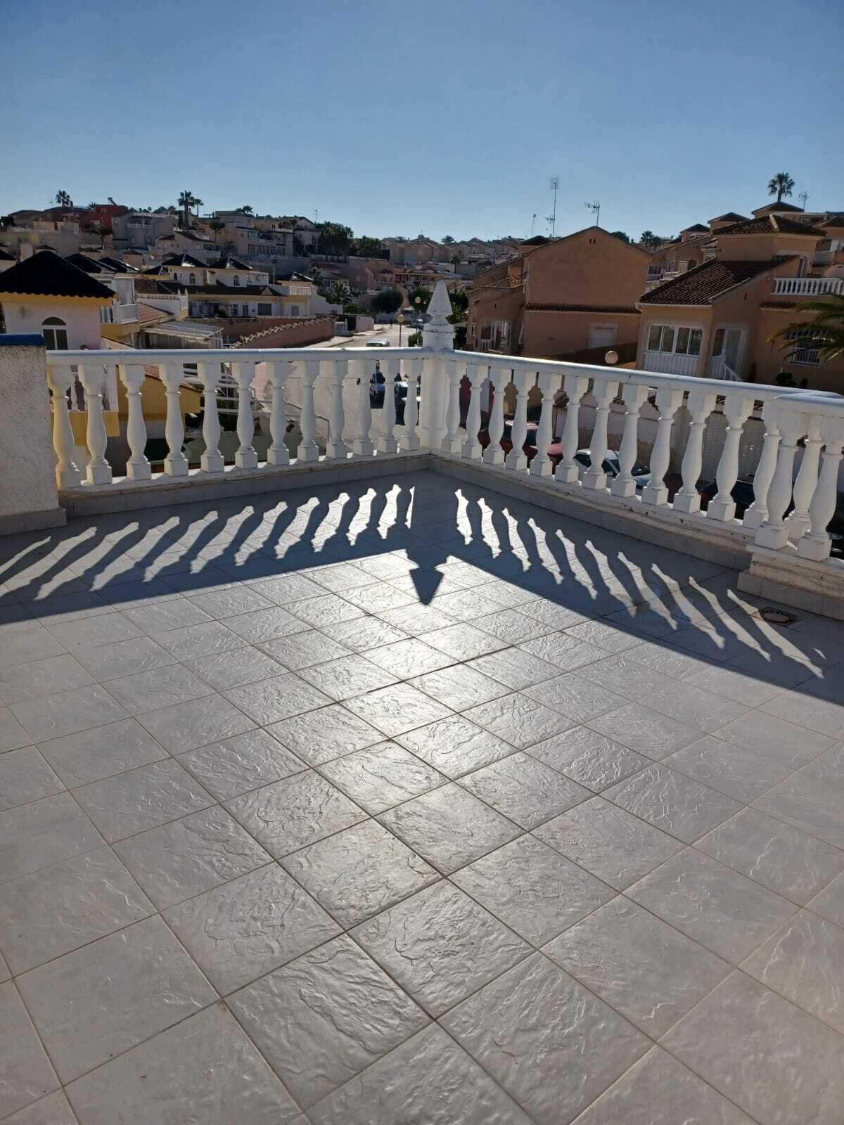 Een betegeld balkon met een witte balustrade, met uitzicht op een woonwijk met huizen en een heldere lucht.