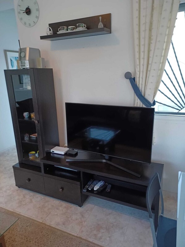 A living room with a flat-screen TV on a dark wooden stand, a small shelf with decorative items, and a clock on the wall. - Exclusive Villa With Private Pool
(Ciudada Quesada)