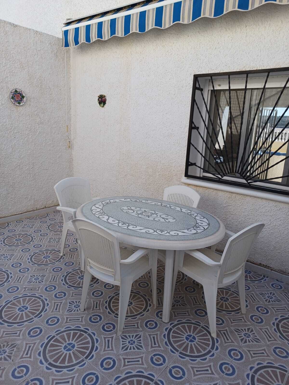 A round table with four chairs, a patterned floor, a striped awning, and a window with a metal grille.