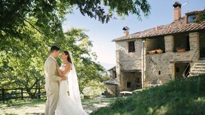 Una coppia si abbraccia in un ambiente rustico all'aperto, con edifici in pietra e vegetazione.