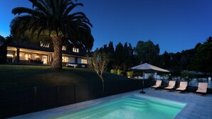 A swimming pool with lounge chairs and an umbrella, surrounded by a landscaped garden and a house with illuminated windows. - Elegant country house with pool. Between Oviedo and Gijón (Siero)
