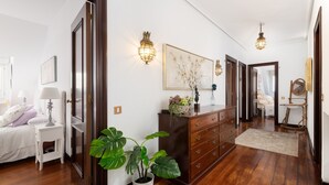 A well-lit hallway with wooden flooring, a white bed with a nightstand, a dresser with drawers, and a potted plant. - Elegant country house with pool. Between Oviedo and Gijón (Siero)
