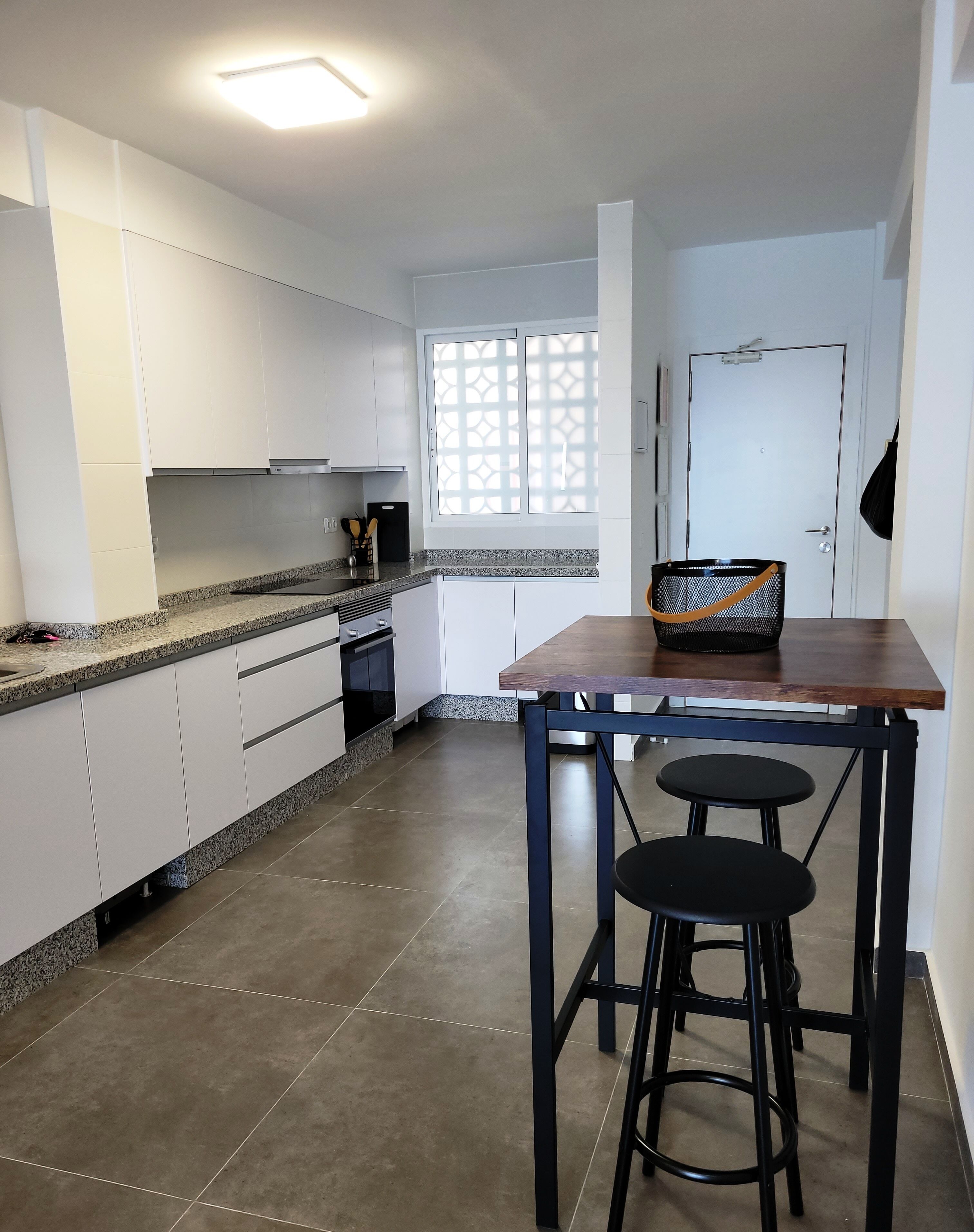Een moderne keuken met witte kastjes, een granieten aanrecht, een houten tafel met twee zwarte stoelen, en een mand op tafel.