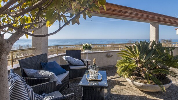 Une terrasse meublée de mobilier en osier, une table avec une bouteille et des verres, et une vue sur la mer.