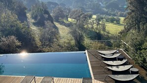 Una piscina infinita con terraza de madera y sillas reclinables con vistas a un paisaje pintoresco con colinas y árboles.