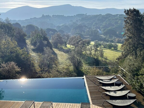 An infinity pool with a wooden deck and lounge chairs overlooking a scenic landscape with hills and trees.
