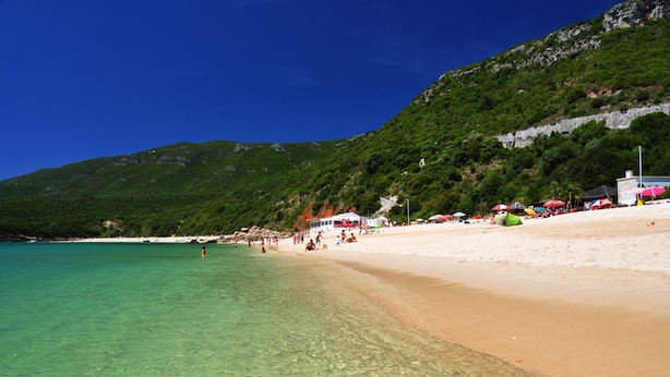 Una playa con aguas turquesas cristalinas, orilla de arena y montañas al fondo.