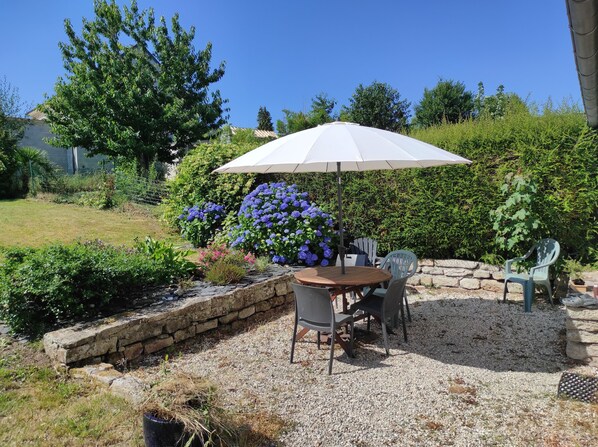 An outdoor patio with a round table, two chairs, and a white umbrella. - Charming penty, 3* gite, enclosed garden with trees (Saint-Évarzec)