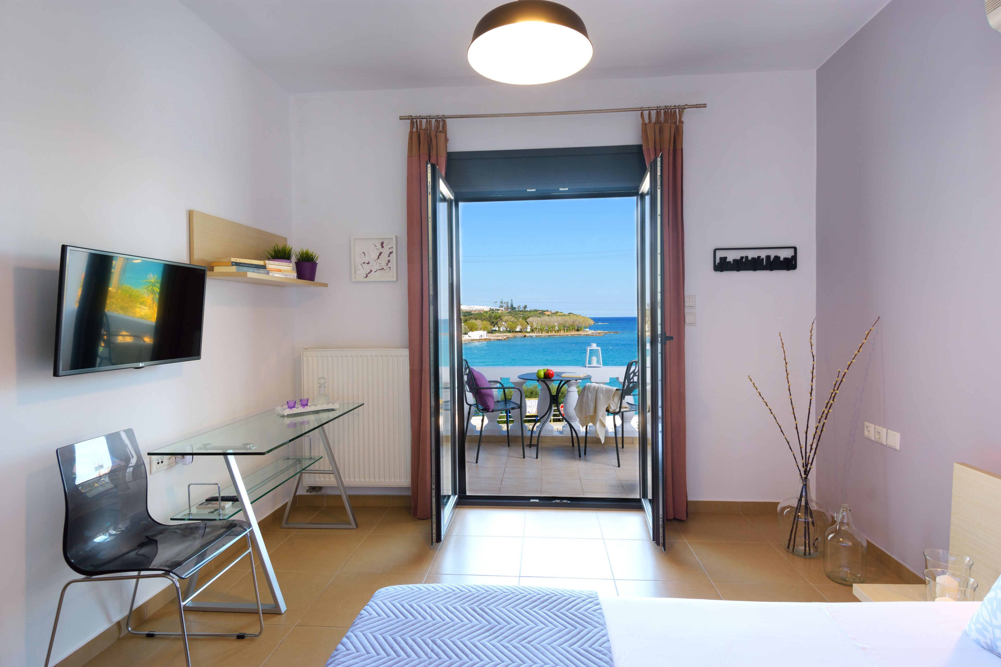 A modern hotel room with a glass desk, a bed, a TV, and a balcony with a view of the sea.