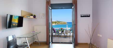 A modern hotel room with a glass desk, a bed, a TV, and a balcony with a view of the sea.