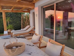 A covered outdoor seating area with lounge chairs, a table, and a large glass door leading to a garden.