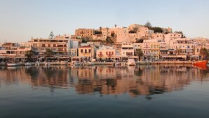 Une ville située au bord de l’eau, avec des bâtiments qui bordent la rive, une étendue d’eau calme où se reflètent les constructions, et une colline imposante sur laquelle se dressent des bâtiments d’architecture historique en arrière-plan.