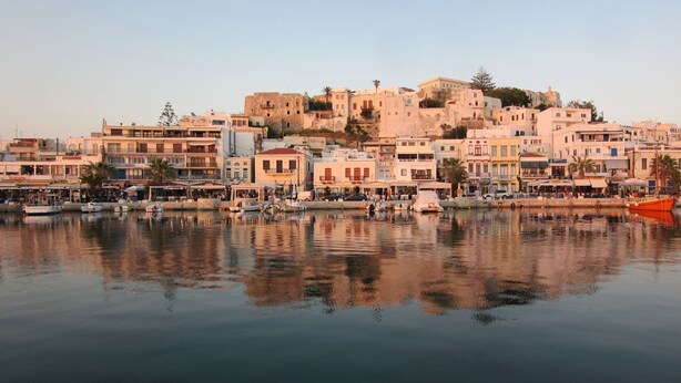 A waterfront town with buildings along the shore, a calm body of water reflecting the structures, and a prominent hill with historical architecture in the background.