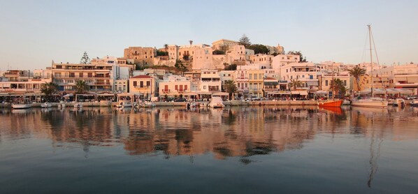 A waterfront town with buildings along the shore, a calm body of water reflecting the structures, and a prominent hill with historical architecture in the background.