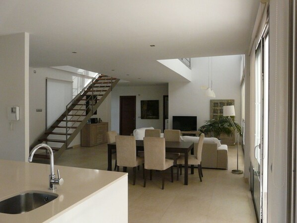 Een moderne keuken met een inbouwspoelbak, een eethoek met een tafel en stoelen, een trap naar een hoger niveau, en een zithoek met een bank en een televisie.