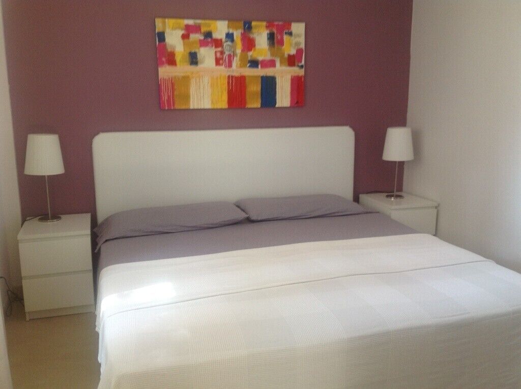 Een slaapkamer met een wit bed, twee nachtkastjes met lampen, en een kleurrijk abstract schilderij aan de muur.