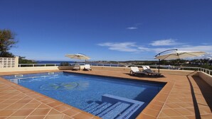 A rectangular swimming pool with tiled edges, surrounded by a tiled deck. There are two white lounge chairs with beige umbrellas, and a view of the sea and distant landscape.