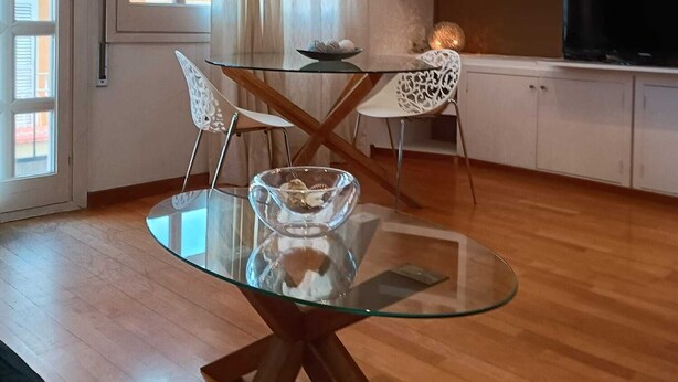 A modern living room with a glass-top dining table, white chairs, a ceiling fan, and a TV on a white cabinet.