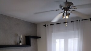 A bedroom with two beds, a ceiling fan, a window with sheer curtains, and a wall-mounted shelf with a potted plant.
