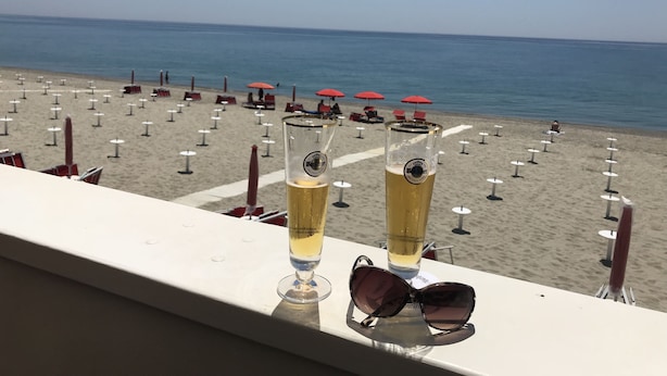 Un balcon avec deux verres de bière et des lunettes de soleil, donnant sur une plage parsemée de parasols et de chaises longues.