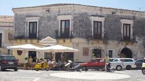 Une place publique pavée de pierres, un bâtiment doté de fenêtres en arc de cercle et des tables installées à l’extérieur, sous des parasols.