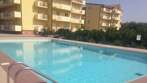 Une piscine dotée d’une rambarde métallique, entourée d’une terrasse carrelée. Au fond, on aperçoit des immeubles jaunes à plusieurs étages, dotés de balcons.