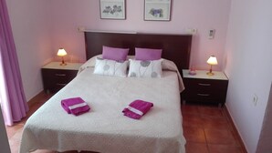 A neatly made bed with a white bedspread and two purple towels, flanked by two bedside tables with lamps, in a room with pink walls and a wooden headboard. - Ground floor with garden and barbecue, 5 minutes from the beach (Alcudia)