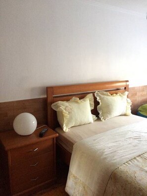 A bedroom with a wooden headboard, two pillows, a bedside table with a remote control, and a white blanket.