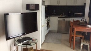 Ein kompakter Wohnraum mit Flachbildfernseher, einem kleinen Glastisch, einem Holzessttisch mit Stühlen und einer Küchenzeile mit weißen Schränken und einem Edelstahl-Geschirrspüler.