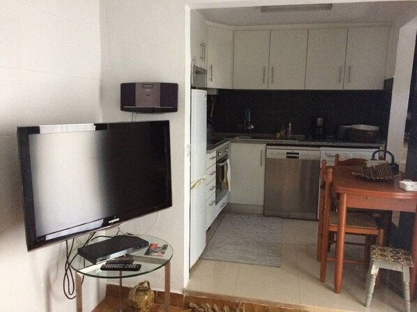 A compact living space with a flat-screen TV, a small glass table, a wooden dining table with chairs, and a kitchenette with white cabinets and a stainless steel dishwasher.